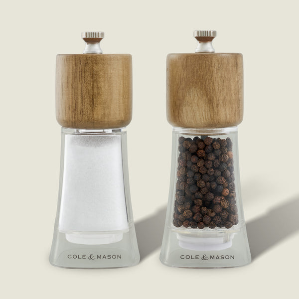 The Cole & Mason Woodbury Salt & Pepper Mill Set includes two clear glass grinders with wooden tops, adjustable coarseness, and adds rustic charm to your table. They stand together on a light background.