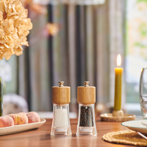 The Cole & Mason Woodbury Salt & Pepper Mill Set, with wooden tops and adjustable coarseness, adds rustic charm to your table by the window amid peaches, a wine glass, dried flowers, and a glowing candle.
