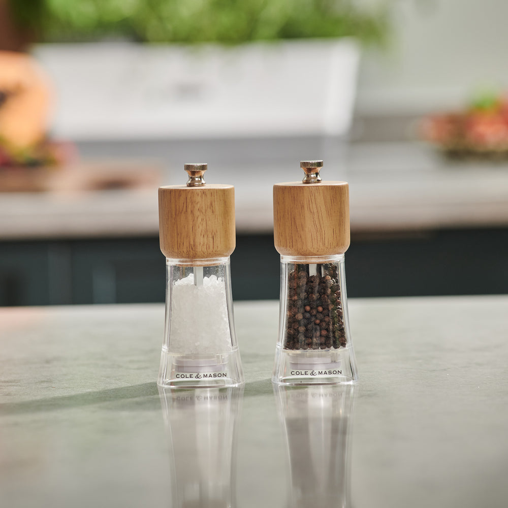 The Cole & Mason Woodbury Salt & Pepper Mill Set features two clear mills with wooden tops, one for salt and one for pepper. Both mills provide adjustable coarseness, all set against a light background for rustic appeal.