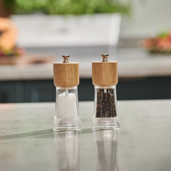 Two clear acrylic grinders with wooden tops from the Cole & Mason Woodbury Salt & Pepper Mill Set sit on a kitchen counter, offering adjustable coarseness and rustic charm and containing coarse salt crystals and whole black peppercorns.