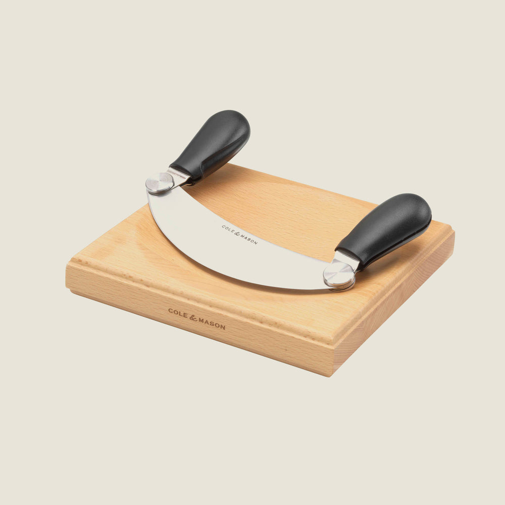 The Cole & Mason UK Woodhall Mezzaluna Hachoir with dual black handles sits on its rectangular chopping board, both branded and ready to enhance your meal prep against a light background.