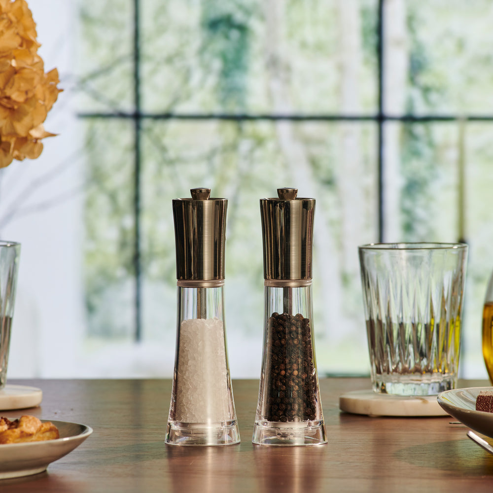 The Cole & Mason Everyday Style Salt & Pepper Mill Set includes two clear glass grinders with stainless steel tops and adjustable settings—ideal for any kitchen. One holds salt, the other peppercorns, displayed side by side on a light background.