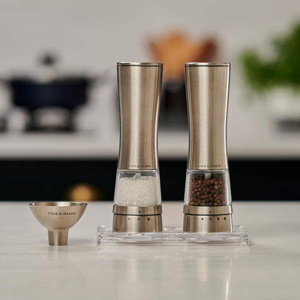 Two Cole & Mason Electric Derwent Bundle salt and pepper grinders stand on a counter, accompanied by a small matching measuring cup. Blurred kitchen items are visible in the background.