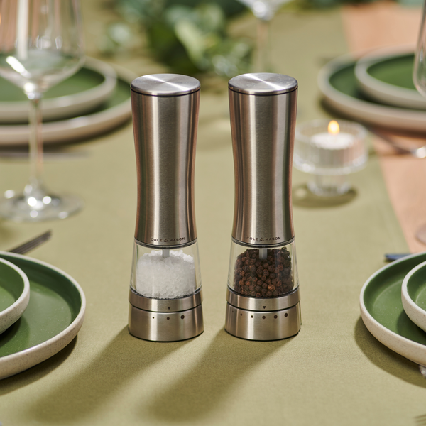 Two Cole & Mason Electric Derwent Salt & Pepper Mills in stainless steel sit side by side on a green tablecloth, surrounded by green plates, glasses, and a lit tealight candle in the background. These rechargeable mills from Cole & Mason blend contemporary convenience with sophisticated style, making them an elegant addition to any dining setting.