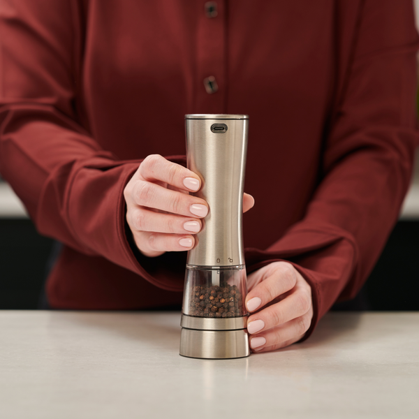 A person in a maroon shirt holds and operates a Cole & Mason Electric Derwent Salt & Pepper Mill, filled with peppercorns, on a light kitchen countertop.