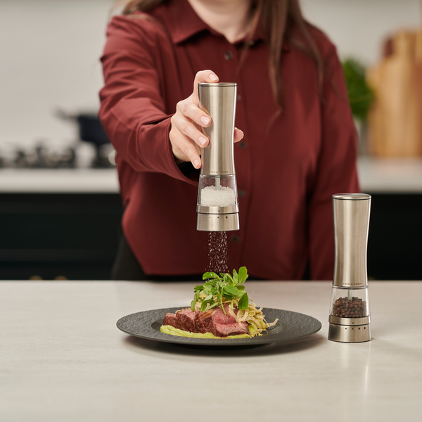 A person in a maroon shirt seasons a plated meal with the Cole & Mason Electric Derwent Salt & Pepper Mill, which sits beside a matching rechargeable grinder on the kitchen counter.