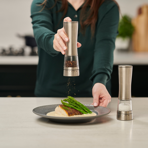 A person uses a Cole & Mason Electric Derwent Salt & Pepper Mill to season a plated dish of fish and asparagus, with another electric salt and pepper mill from the same collection nearby on the kitchen counter.