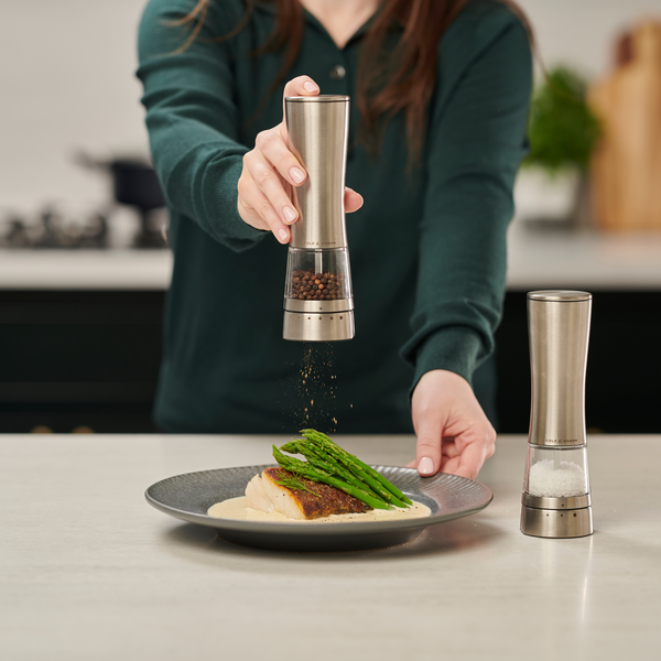 A person uses a Cole & Mason Electric Derwent Salt & Pepper Mill to season a plated dish of fish and asparagus, with another electric salt and pepper mill from the same collection nearby on the kitchen counter.