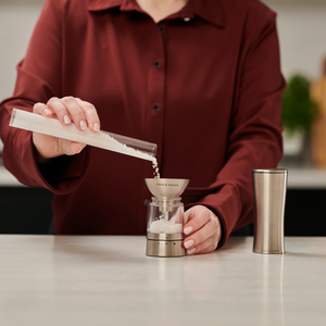 A person in a red shirt pours coarse salt from a clear container into a metal funnel atop a glass salt grinder, which features the sleek design of the Cole & Mason Electric Derwent Salt & Pepper Mills, with a stainless steel cup nearby on the kitchen counter.