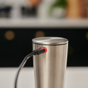 A Cole & Mason Electric Derwent Salt & Pepper Mill is shown charging with a black cable plugged in and a small red light glowing near the port, much like a rechargeable kitchen gadget. The background features blurred kitchen decor.