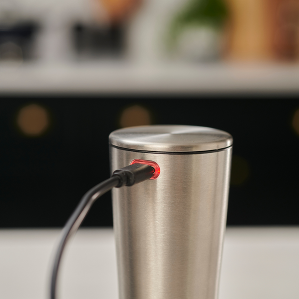 A Cole & Mason Electric Derwent Salt & Pepper Mill is shown charging with a black cable plugged in and a small red light glowing near the port, much like a rechargeable kitchen gadget. The background features blurred kitchen decor.
