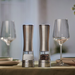 Two Cole & Mason Electric Derwent Salt & Pepper Mills, featuring a sleek stainless steel design, are displayed on a set dining table adorned with plates and wine glasses, illuminated by soft natural lighting in the background.