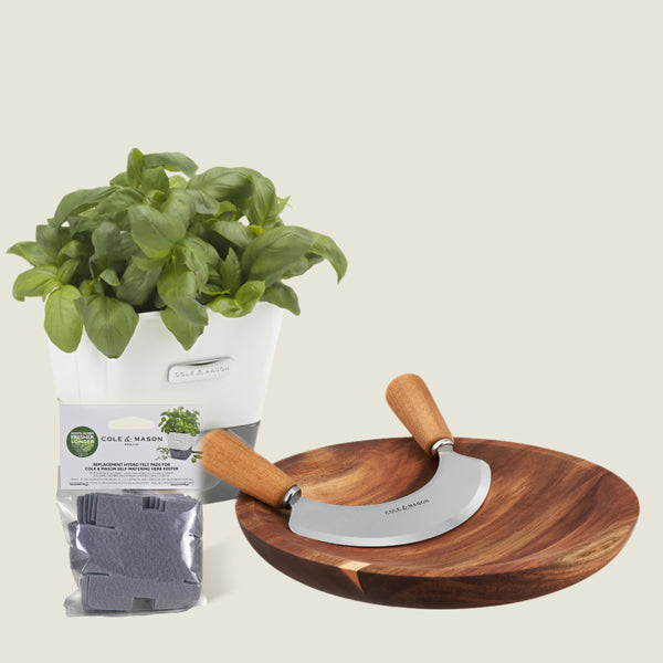 The Cole & Mason Single Herb Keeper, Chopper & Refills Bundle includes a white-potted basil plant, soil pod packet, wooden bowl, and stainless steel Mezzaluna chopper—everything you need to prepare fresh herbs with ease.