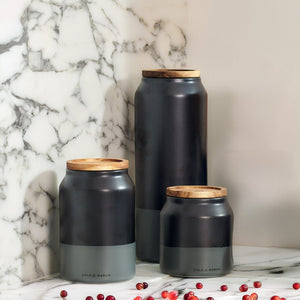 Three matte black and gray Cole & Mason Hinxton Ceramic Storage Jars with wooden lids are arranged on a white marble surface, with small red berries scattered nearby.