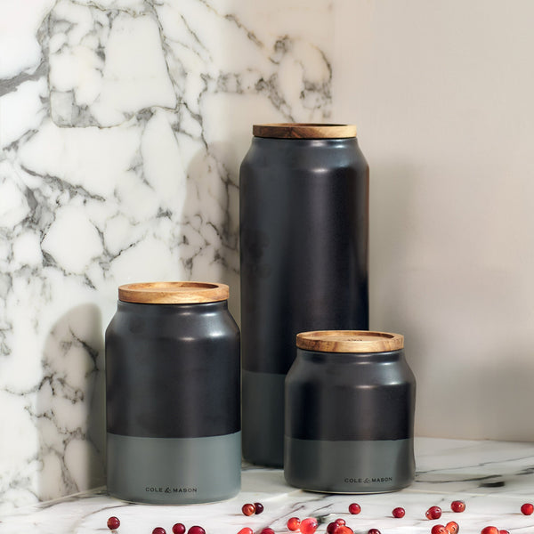 Three matte black and gray Cole & Mason Hinxton Ceramic Storage Jars with wooden lids are arranged on a white marble surface, with small red berries scattered nearby.