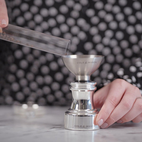 A person in a black and white polka dot shirt fills a stainless steel spice grinder with pepper using a small clear tube. The grinder sits on a marble surface and is labeled Cole & Mason.