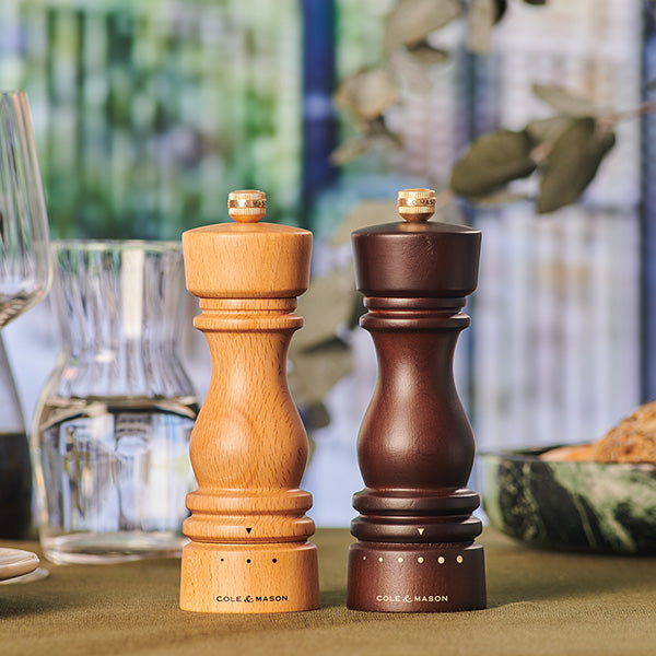 The Fast Bundle London Grind Select Salt & Pepper Mill Bundle—one light beech wood, one dark—stands side by side on a green tablecloth with glassware, plates, and a leafy background in soft focus.