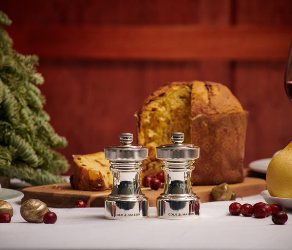 Two silver Cole & Mason salt and pepper grinders sit on a white tablecloth, with cranberries, nuts, a Christmas tree branch, and a sliced panettone in the background.