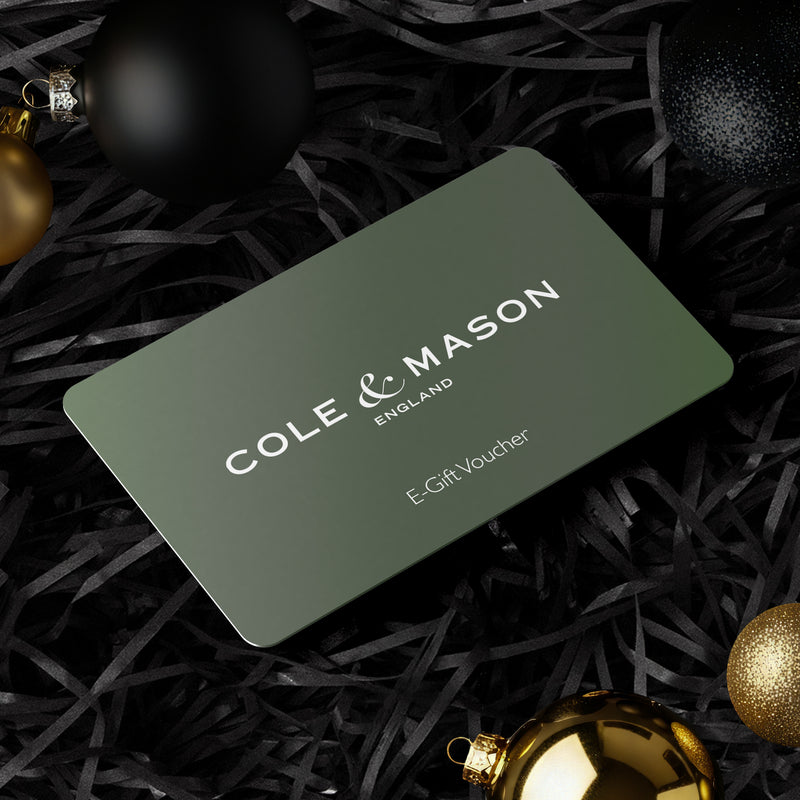 A Cole & Mason UK e-Gift Voucher lies on black shredded paper, surrounded by black and gold Christmas baubles—the ideal gift for anyone who loves luxury kitchenware or premium salt and pepper sets.