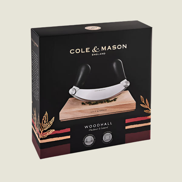 The Cole & Mason UK Woodhall Mezzaluna Hachoir with dual black handles sits on its rectangular chopping board, both branded and ready to enhance your meal prep against a light background.